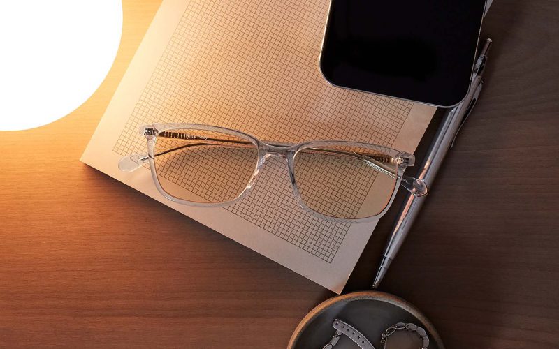 Clear rectangular eyeglasses on a note pad with a light, phone, and pen on a table